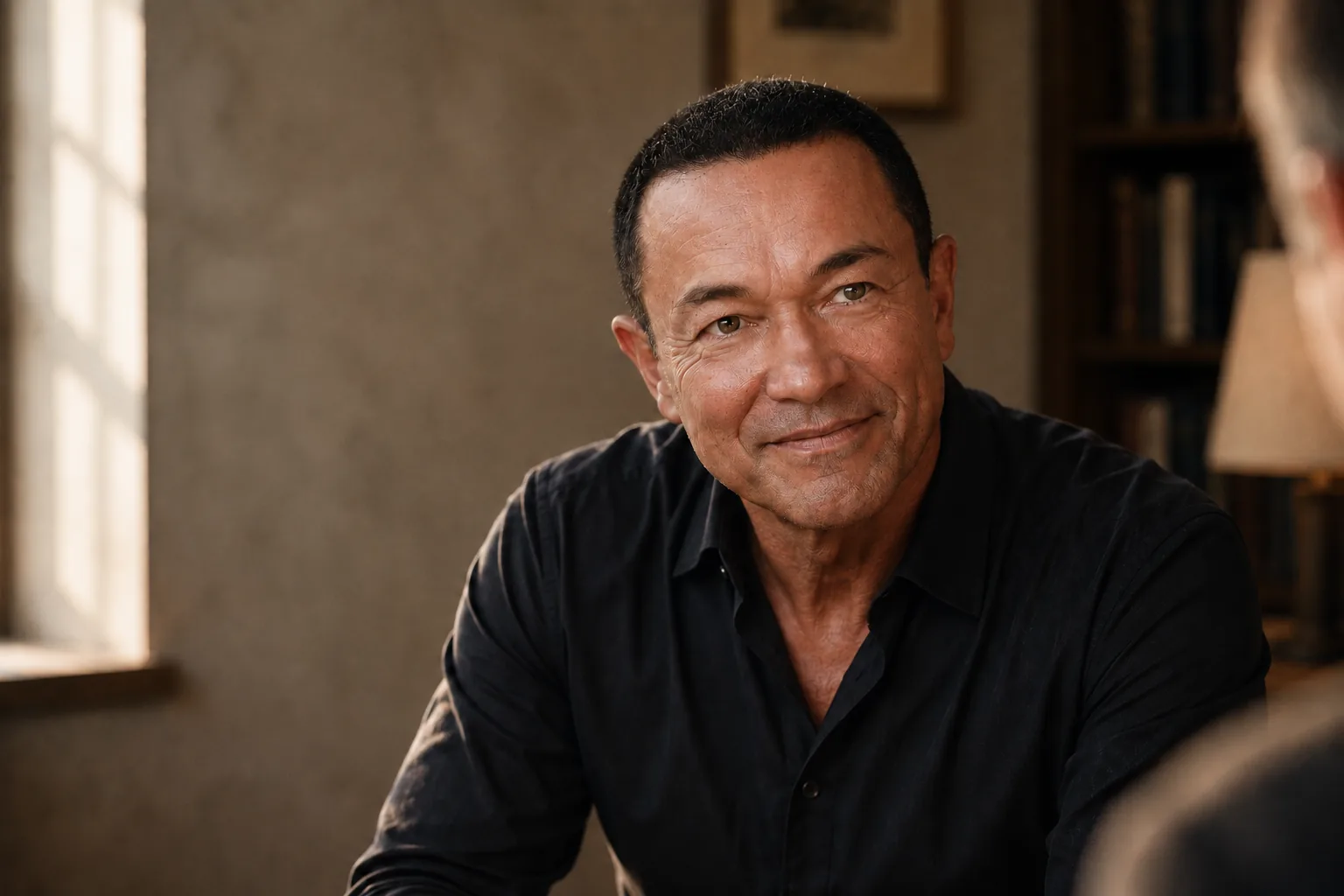 A coach in a dark shirt, smiling mid-conversation, seated by a window in warm light
